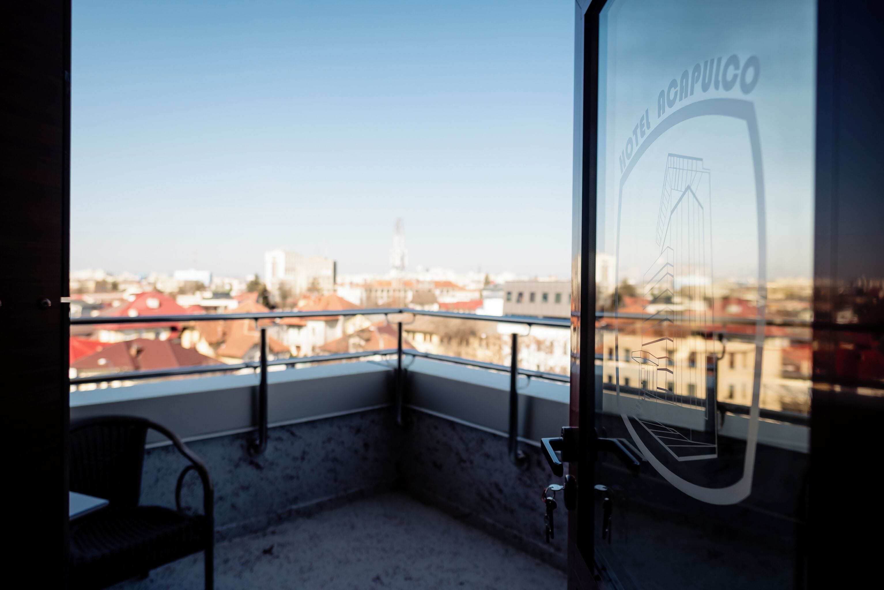 A balcony with a view of a cityscape, a chair, and a glass door with a logo.
