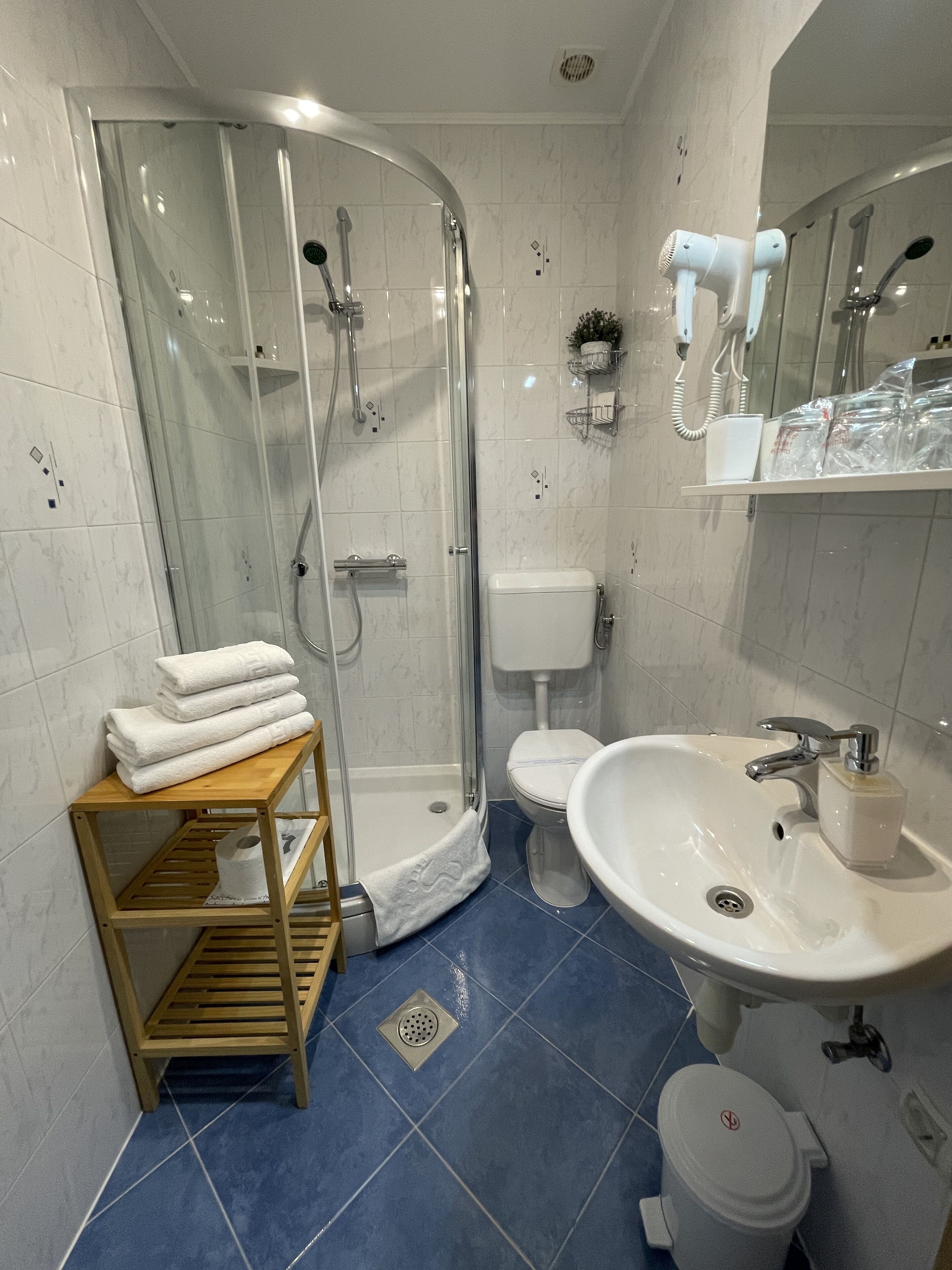 A modern bathroom with a glass-enclosed shower, a white toilet, a sink with a soap dispenser, and a wooden shelf with folded towels.