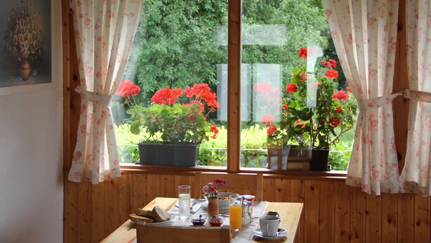 Un comedor con una mesa dispuesta para el desayuno, una ventana con cortinas, y vistas a flores rojas y follaje verde en el exterior.