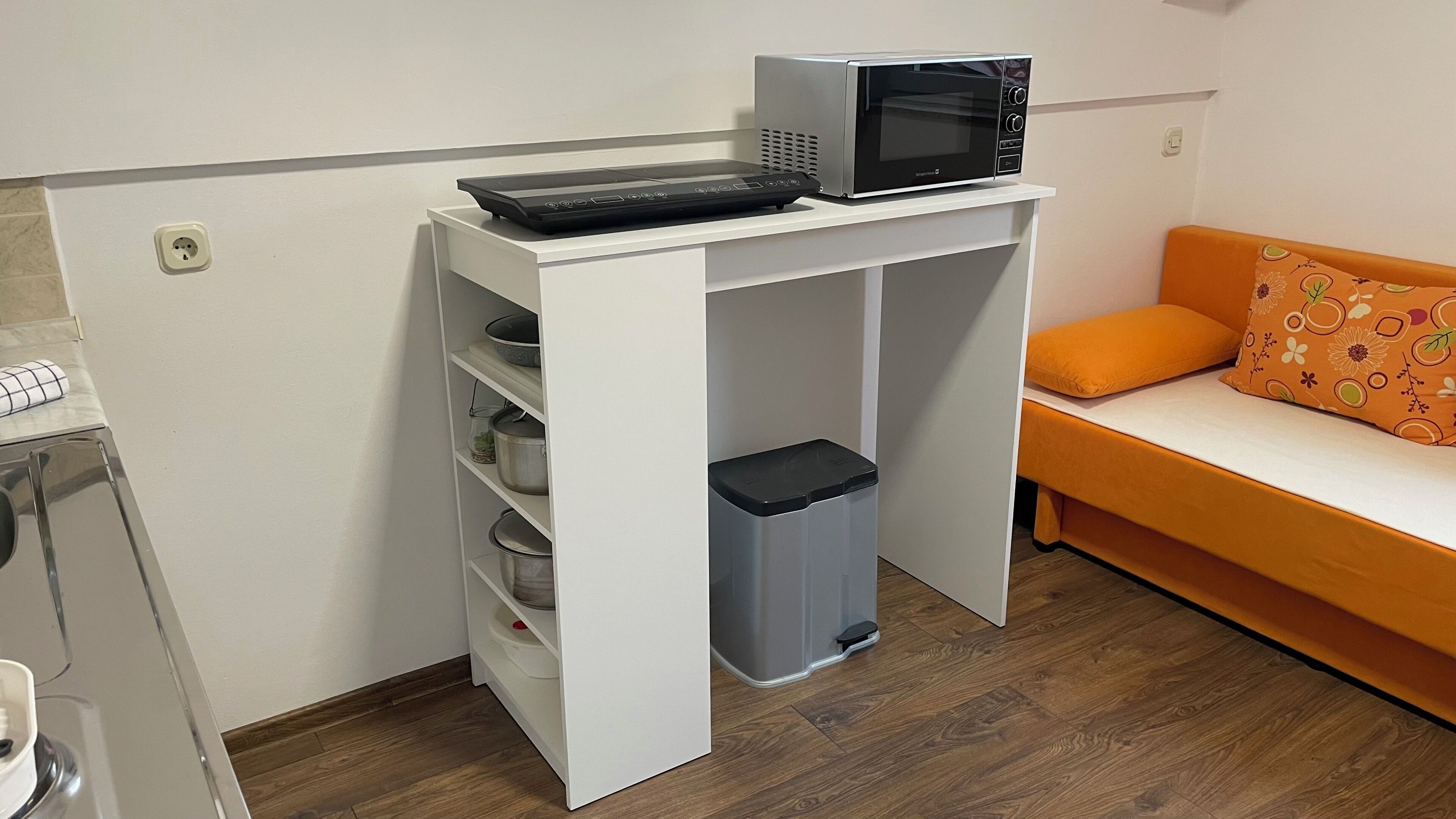 A compact room with a kitchenette, a microwave, a small trash bin, and a sofa with orange cushions.