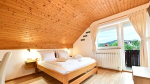 Un dormitorio con techo de madera, cama con ropa blanca, mesita de noche, escritorio y ventana con vista a la vegetación.