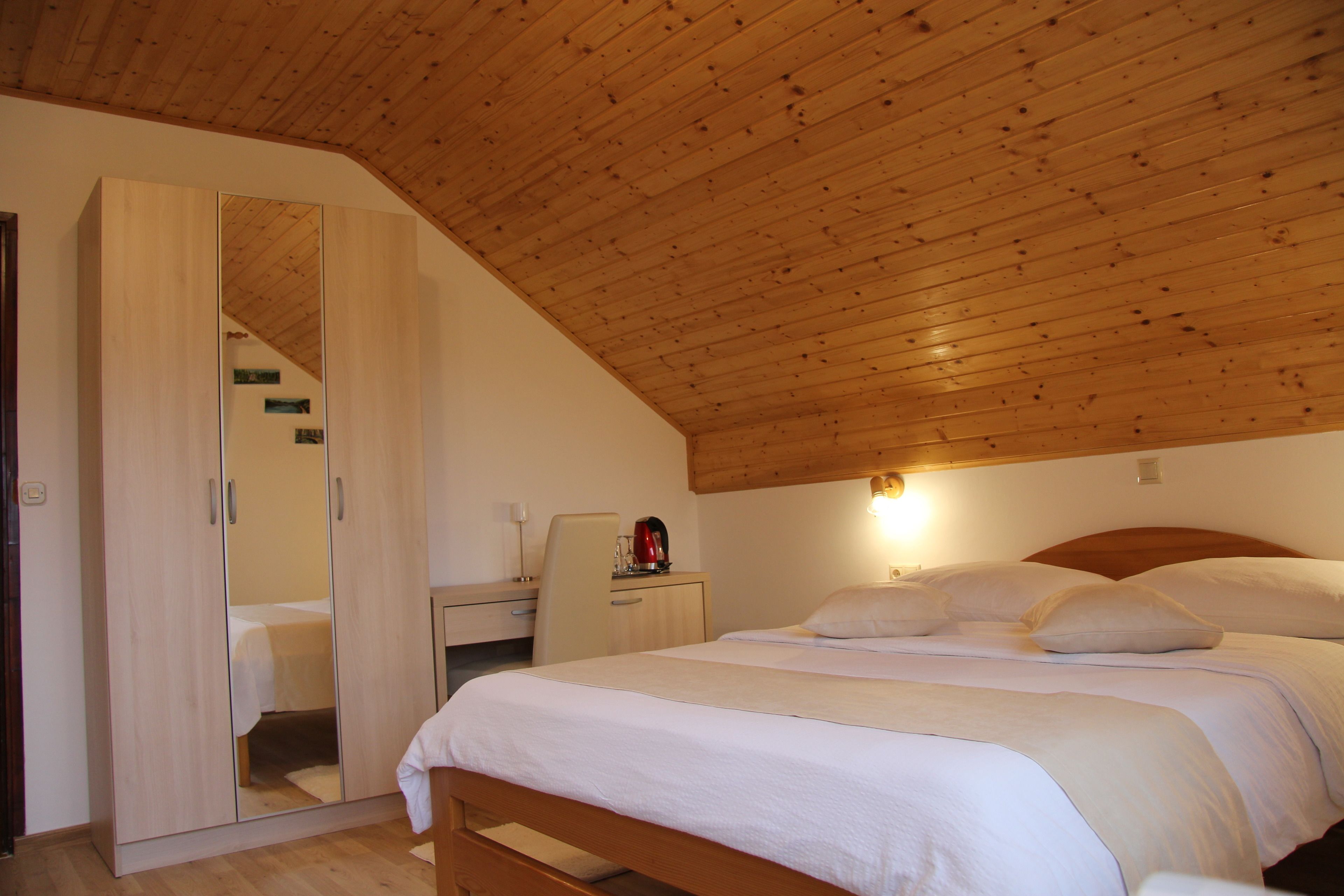A bedroom with a wooden ceiling, a bed with white bedding, a nightstand with a red kettle, and a wardrobe with a mirror.