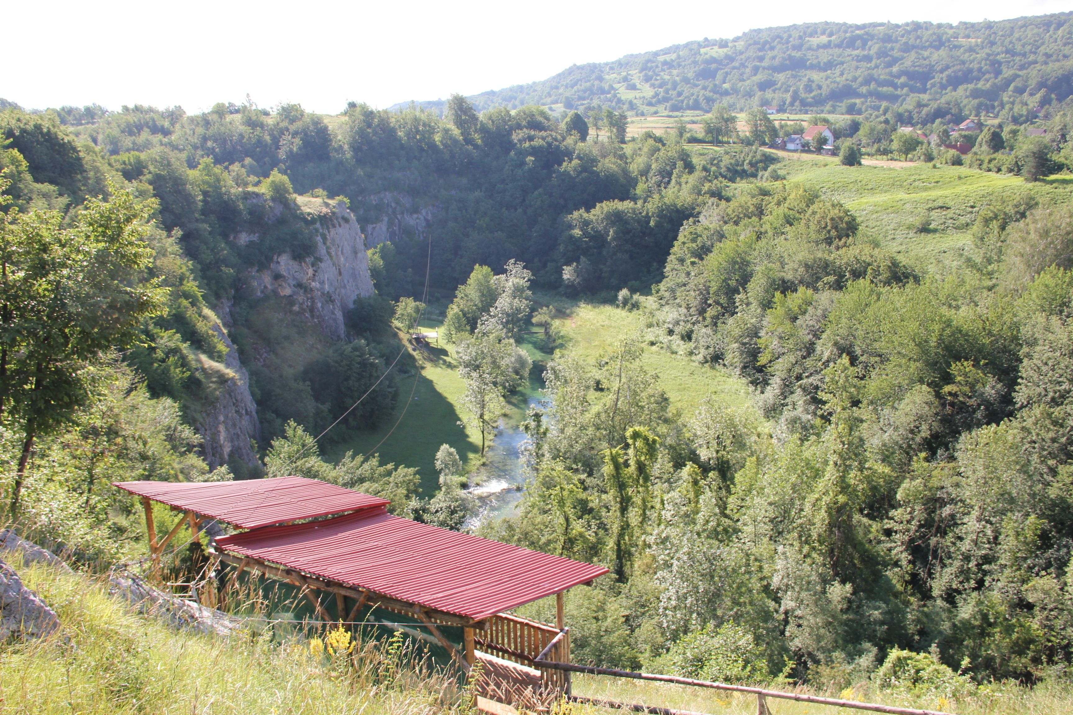 A scenic view of a valley with a river, a wooden structure with a red roof, and lush greenery.
