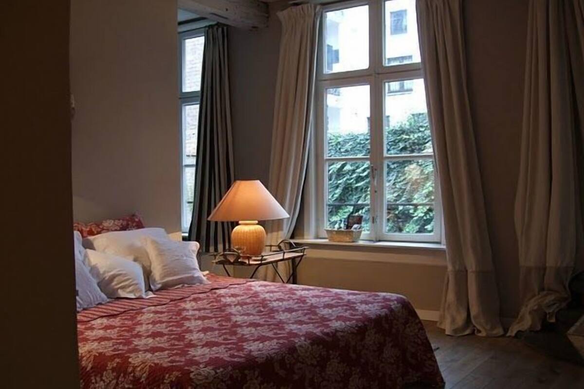 A bedroom with a bed, a bedside table with a lamp, and a window with curtains.