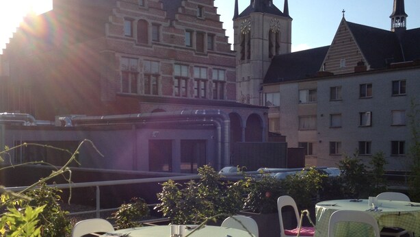 An outdoor seating area with tables and chairs, overlooking a historic building with multiple spires. - Corbie Geel (Geel)