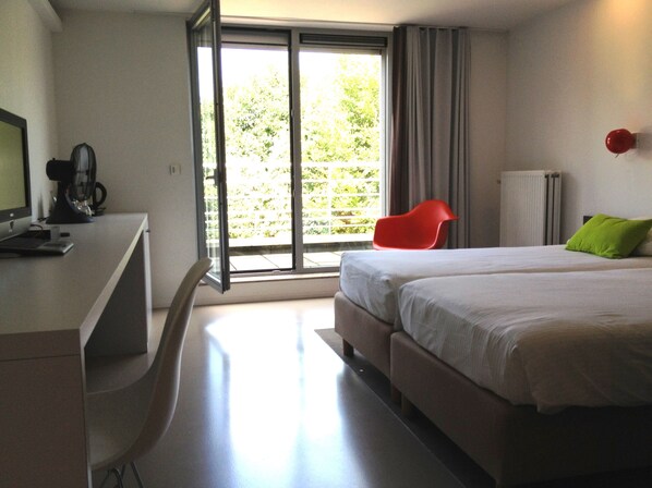 A modern hotel room with a large bed, a desk with a computer, a red chair, and a view through the sliding glass door. - Corbie Geel (Geel)