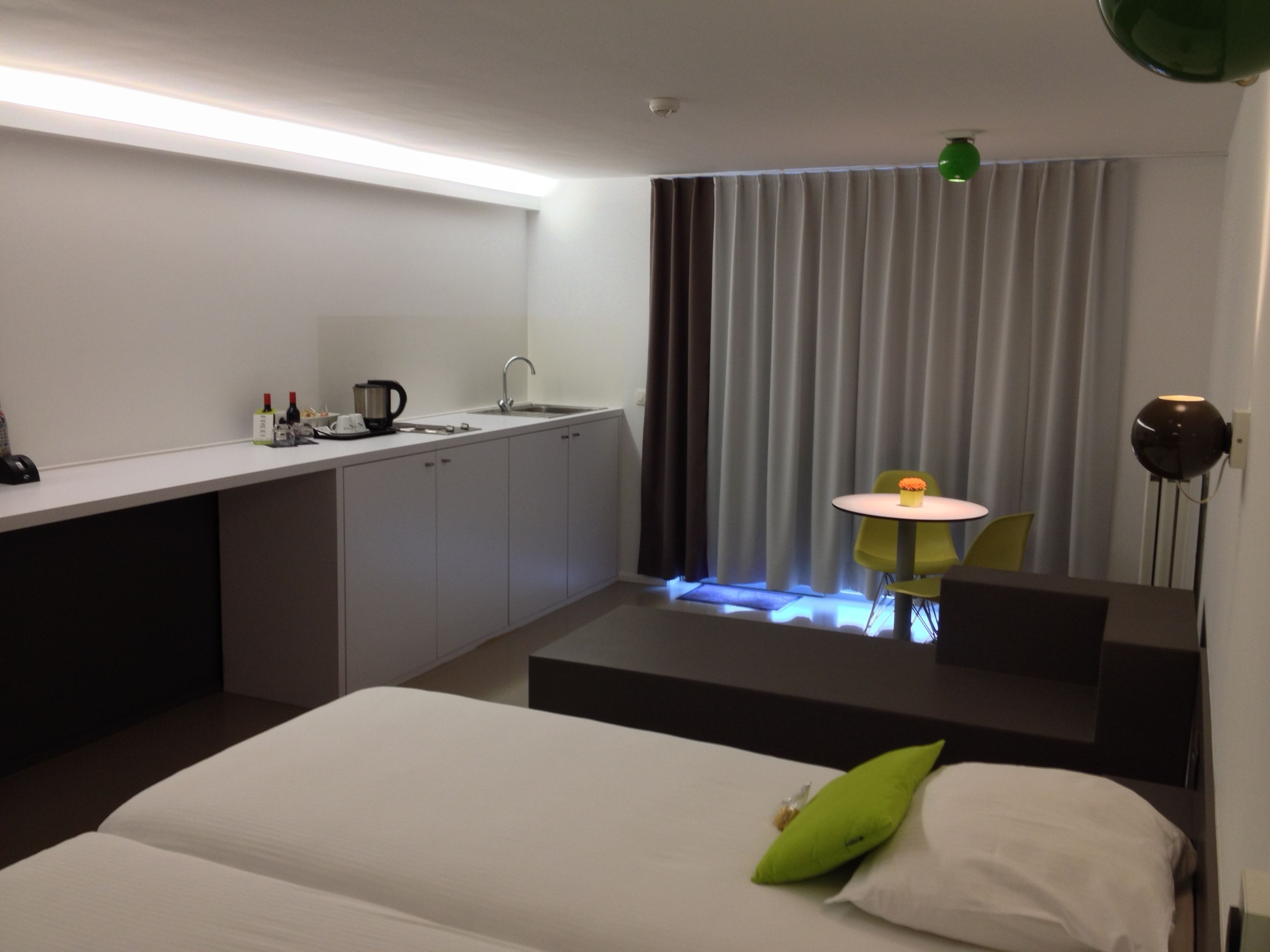 Una habitación de hotel moderna con una cama, un escritorio, una mesita, una silla, un lavamanos y un ventanal con cortinas.