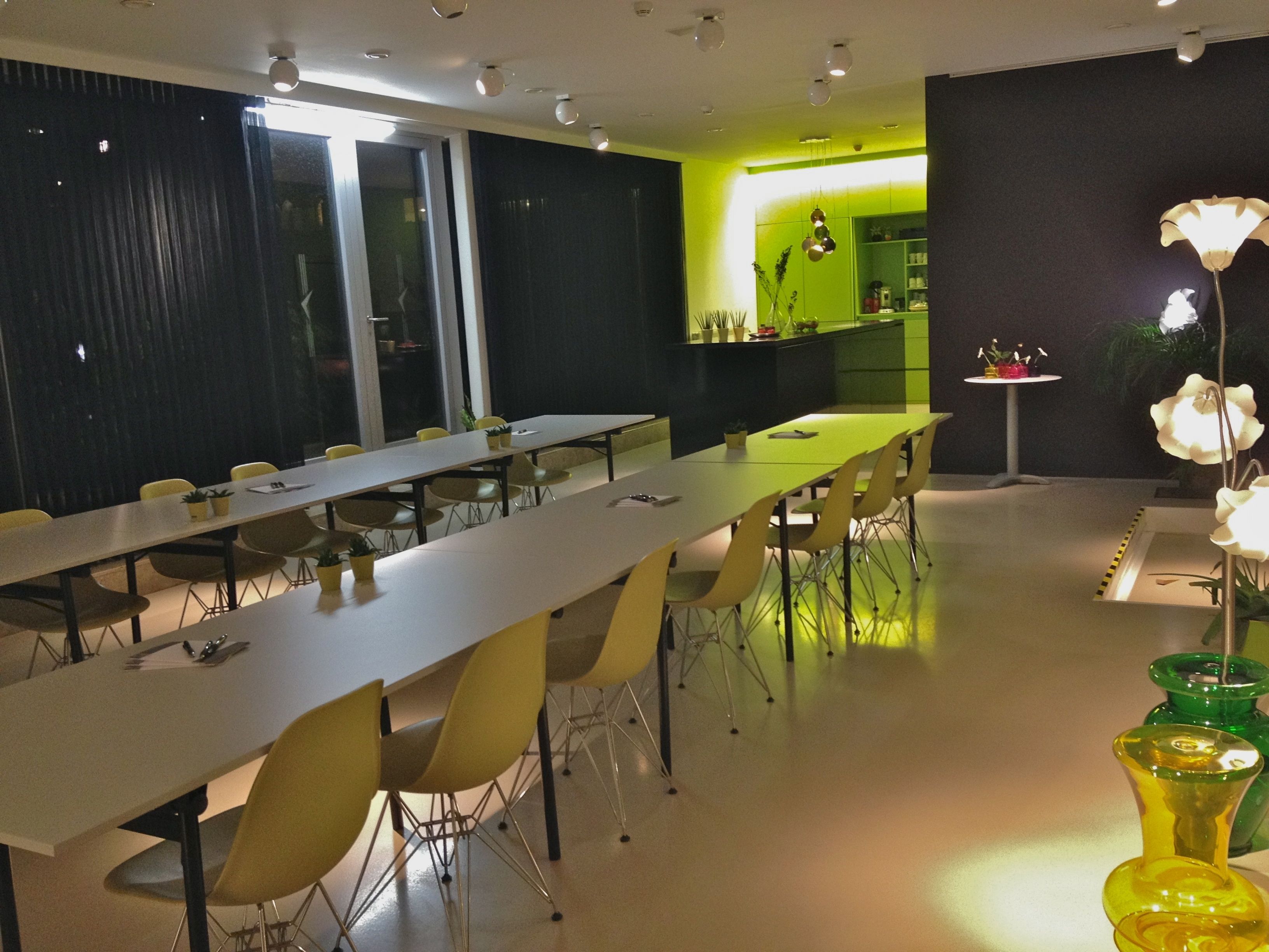 Une salle de conférence moderne, dotée de longues tables et de chaises jaunes, d’un coin cuisine et d’une composition florale décorative.