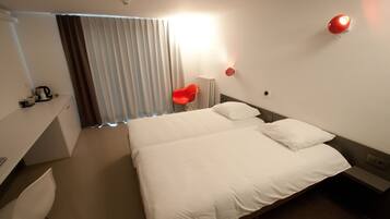 Une chambre d’hôtel avec un grand lit, un bureau avec une chaise, un radiateur et une fenêtre avec des stores.