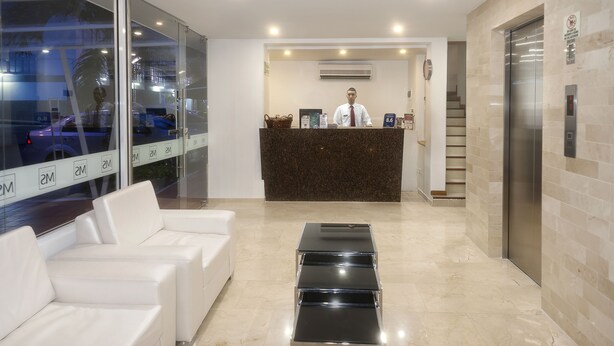 A hotel reception area with a receptionist, a reception desk, a waiting area with white sofas, and an elevator. - Basic Hotel Centenario by Hoteles MS (Cali)