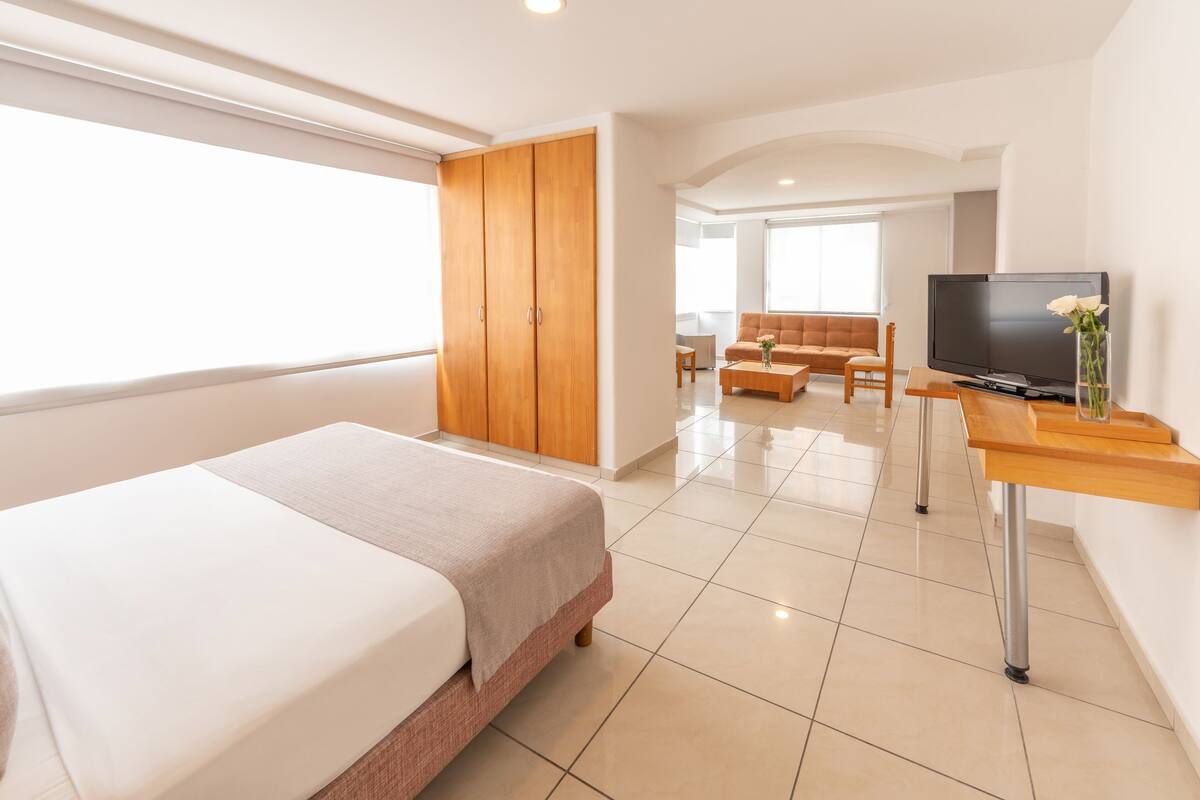 A modern hotel room with a large bed, a TV, a wooden wardrobe, a sofa, and a dining table.