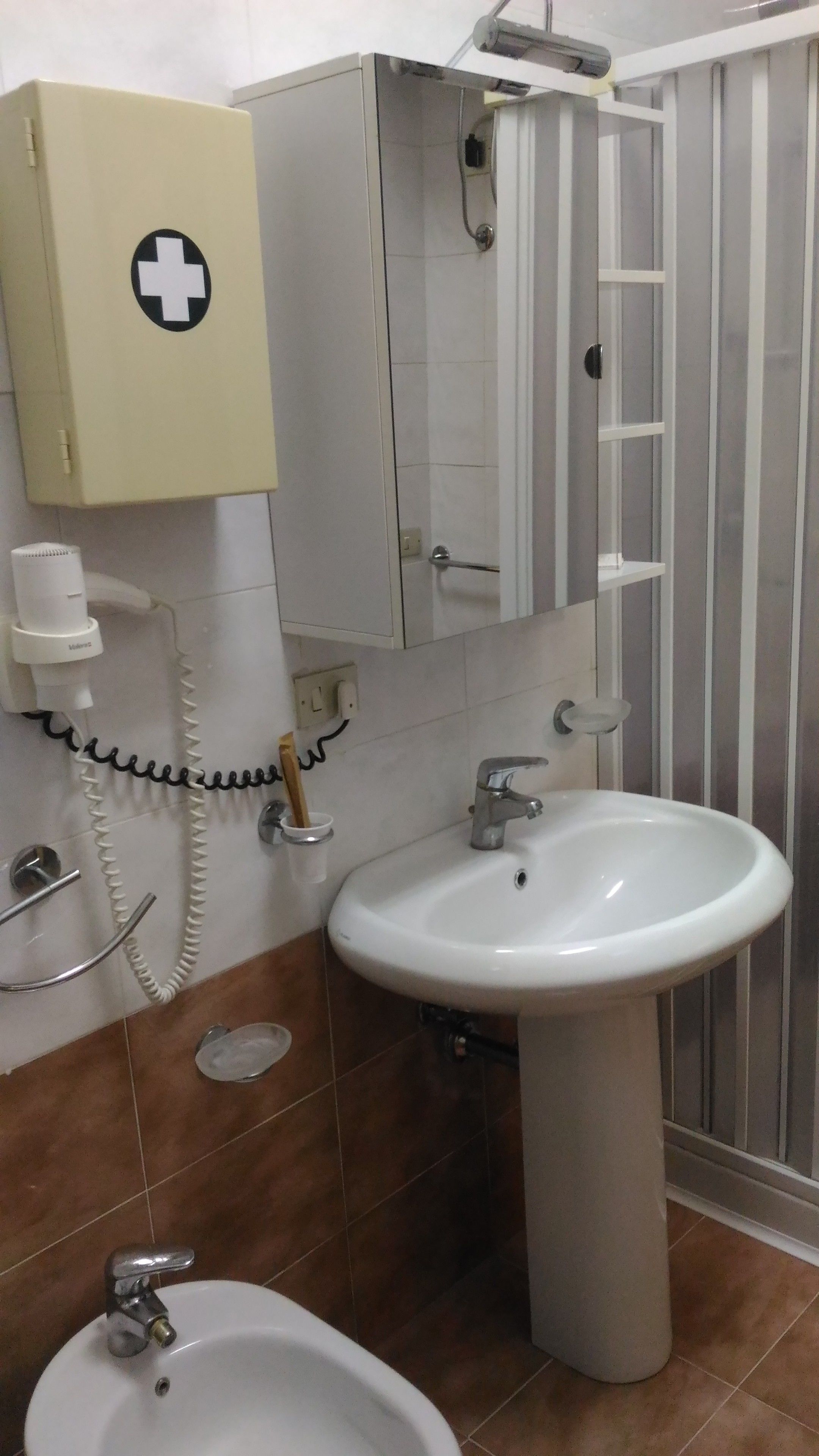 Une salle de bain avec un lavabo blanc, un bidet, des toilettes, un miroir et un meuble orné d’une croix médicale.