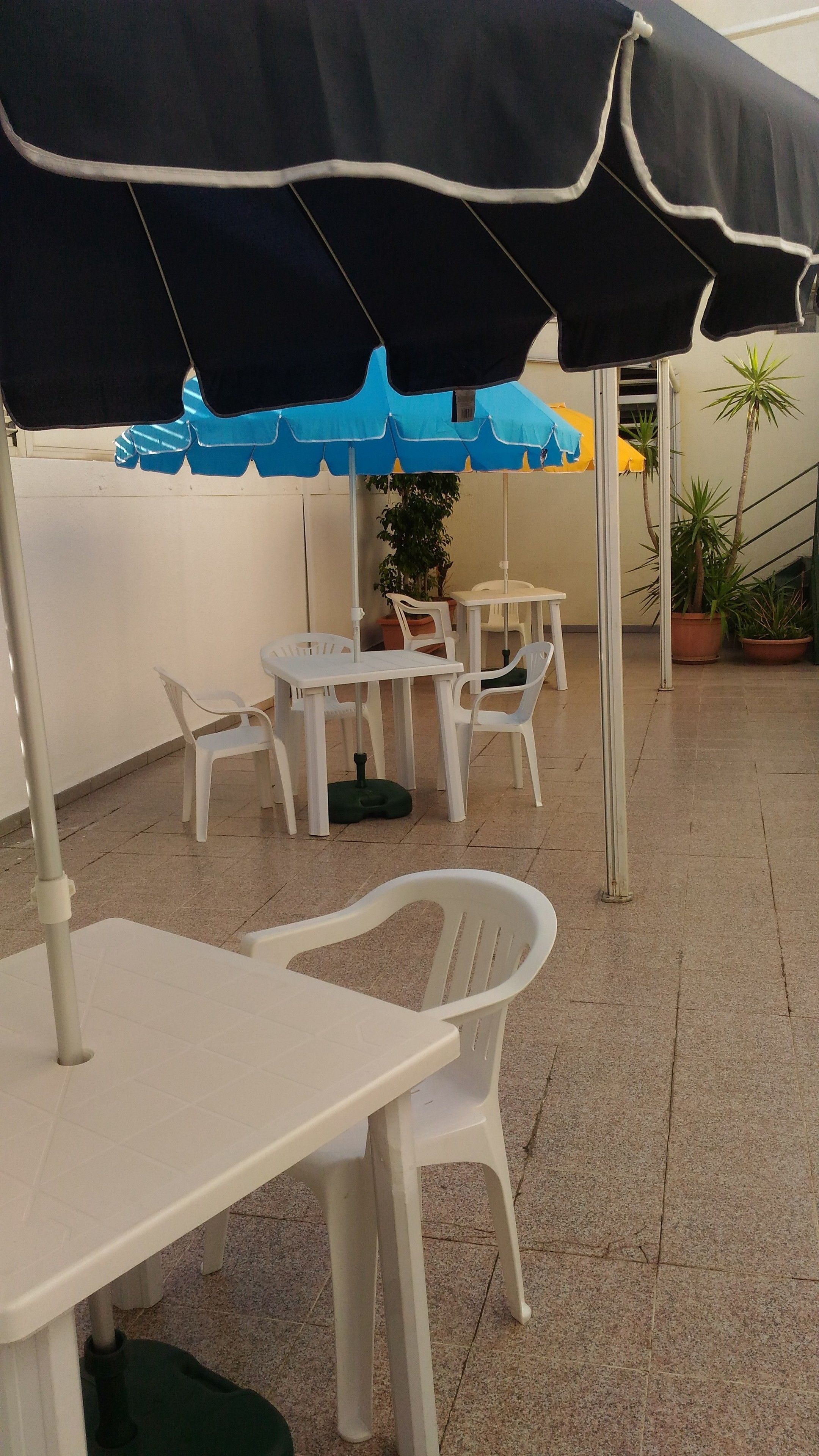 Eine Terrasse mit weißen Plastiktischen und -stühlen, einem schwarz-blauen Sonnenschirm und Topfpflanzen.