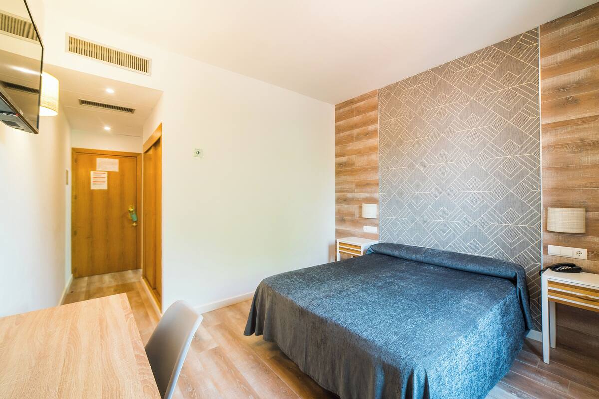 Habitación de hotel con cama, escritorio, silla y pared de madera con diseño geométrico.
