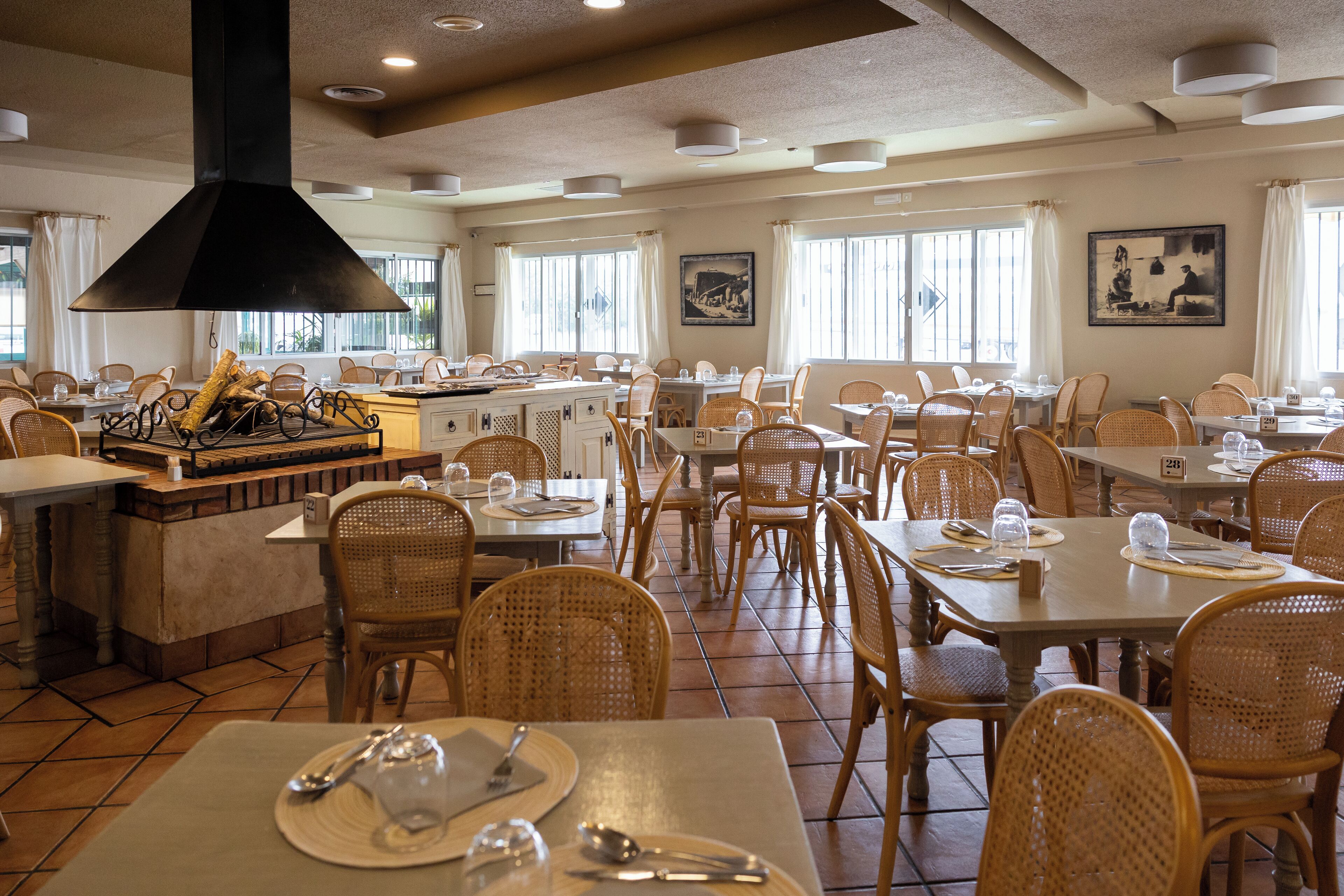 Ein geräumiges Restaurant mit mehreren Esstischen und Korbstühlen, großen Fenstern mit Vorhängen sowie einer zentral gelegenen Küche.