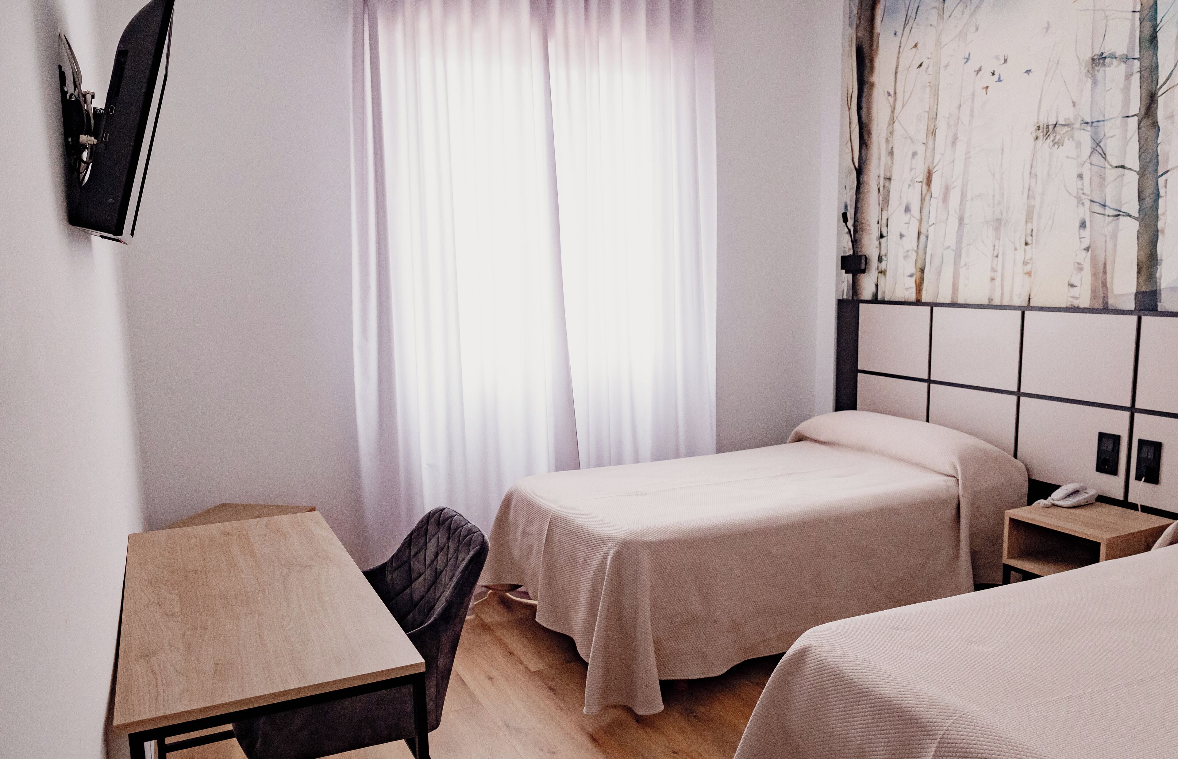 Camera d'albergo con due letti, una scrivania, una sedia, una TV a parete e una finestra con tende.