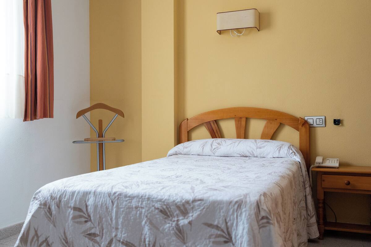 Una cama individual con cabecera de madera, una mesita de noche con teléfono y una lámpara de pared.