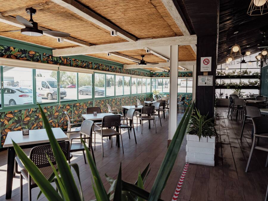 Interior de restaurante con vigas de madera en el techo, ventanas amplias y plantas de interior.