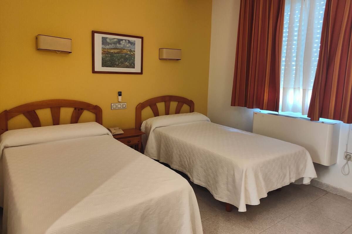 Una habitación con dos camas individuales, un cuadro en la pared, una ventana con cortinas y un termostato en la pared.