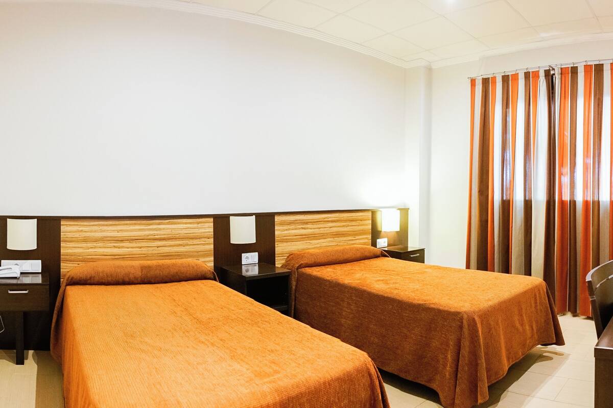 Habitación de hotel con dos camas, un escritorio, una silla, una lámpara y una cortina.