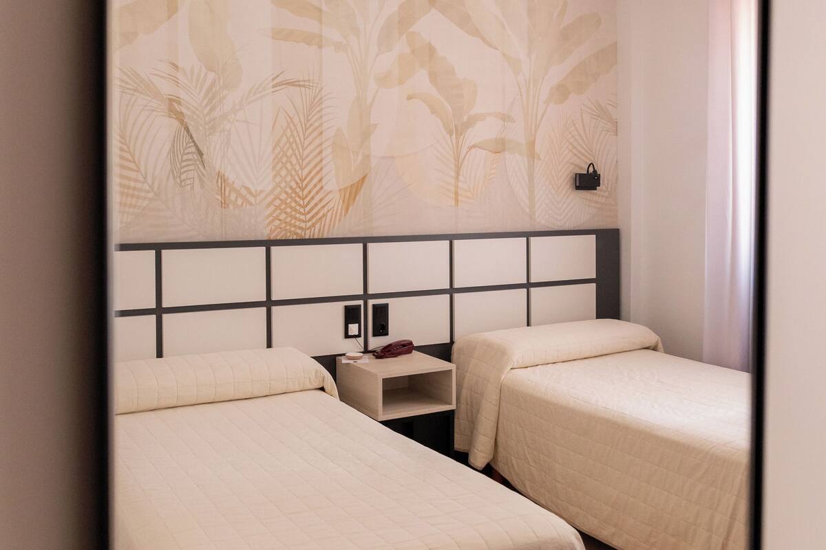 Habitación de hotel con dos camas, una mesita de noche y un cabecero con motivos de hojas.