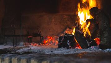 A close-up of a fire with burning logs and embers, with a metal chair and a brick wall in the background.