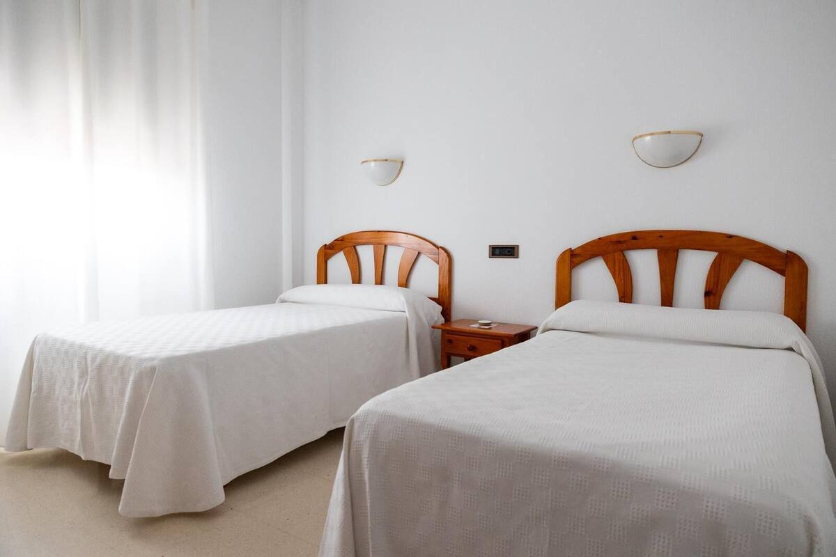Una habitación con dos camas individuales, cabeceros de madera, ropa de cama blanca, una mesita de noche con lámpara y una lámpara fijada a la pared.