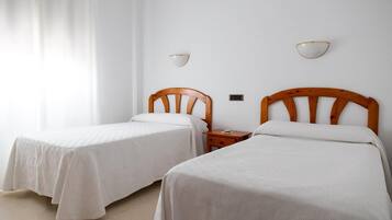 A room with two single beds, wooden headboards, white bedding, a bedside table with a lamp, and a wall-mounted light fixture.