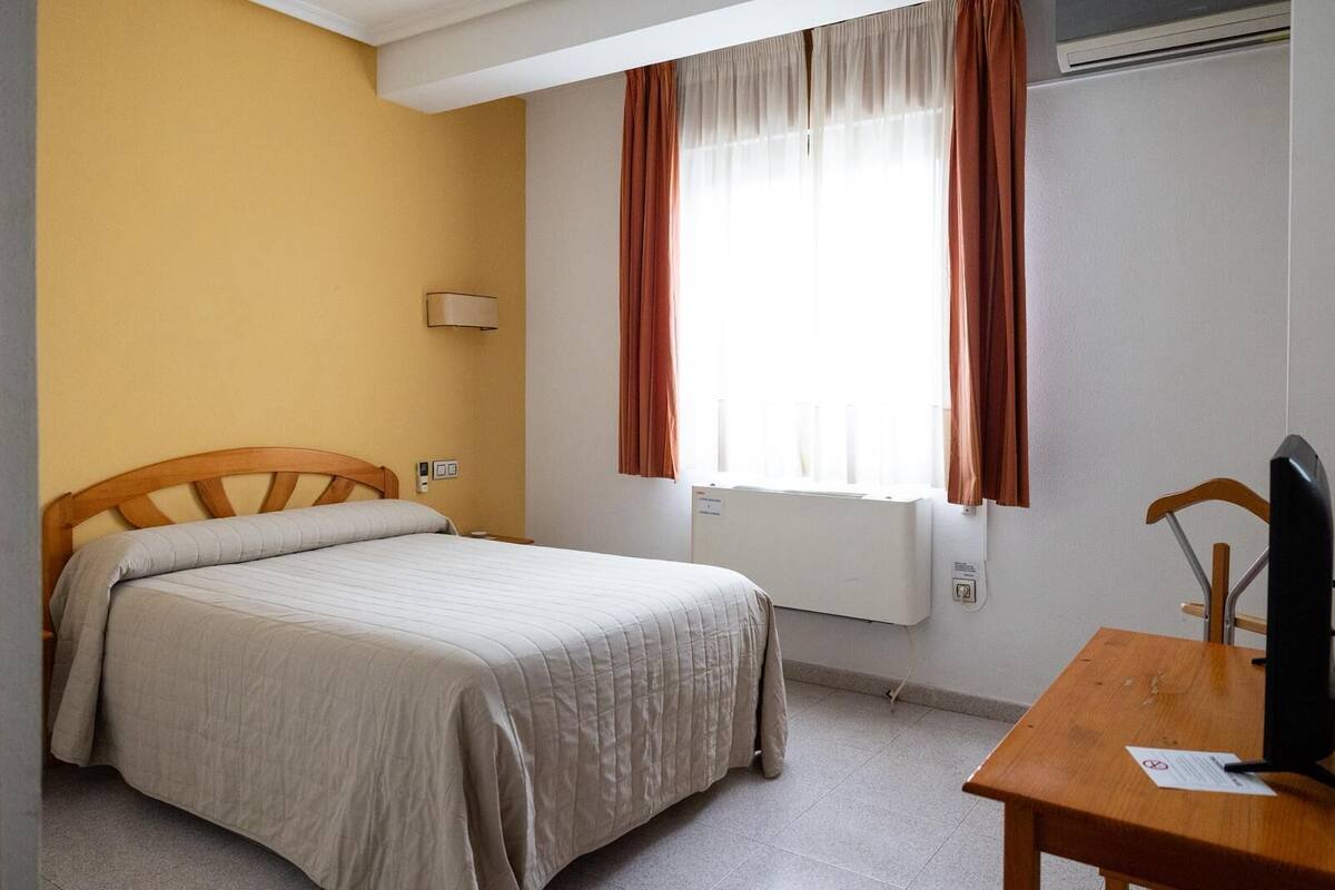 Una cama individual con cabecera de madera, una mesita de noche con televisor y una ventana con cortinas.