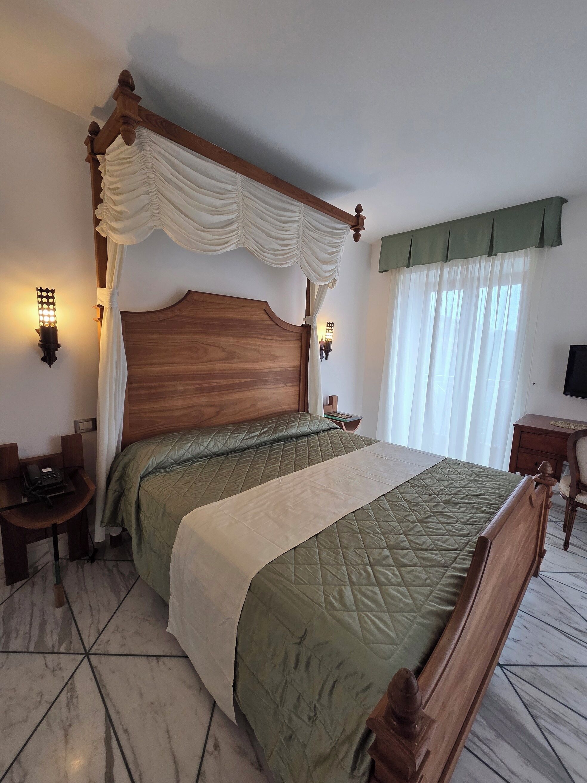 A bedroom with a wooden bed, a nightstand, a desk with a telephone, and a television on a wooden stand.