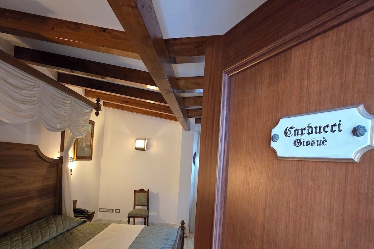 A bedroom with a bed, wooden beams, a sign reading 'Carducci Biosoque', a chair, and a small table.