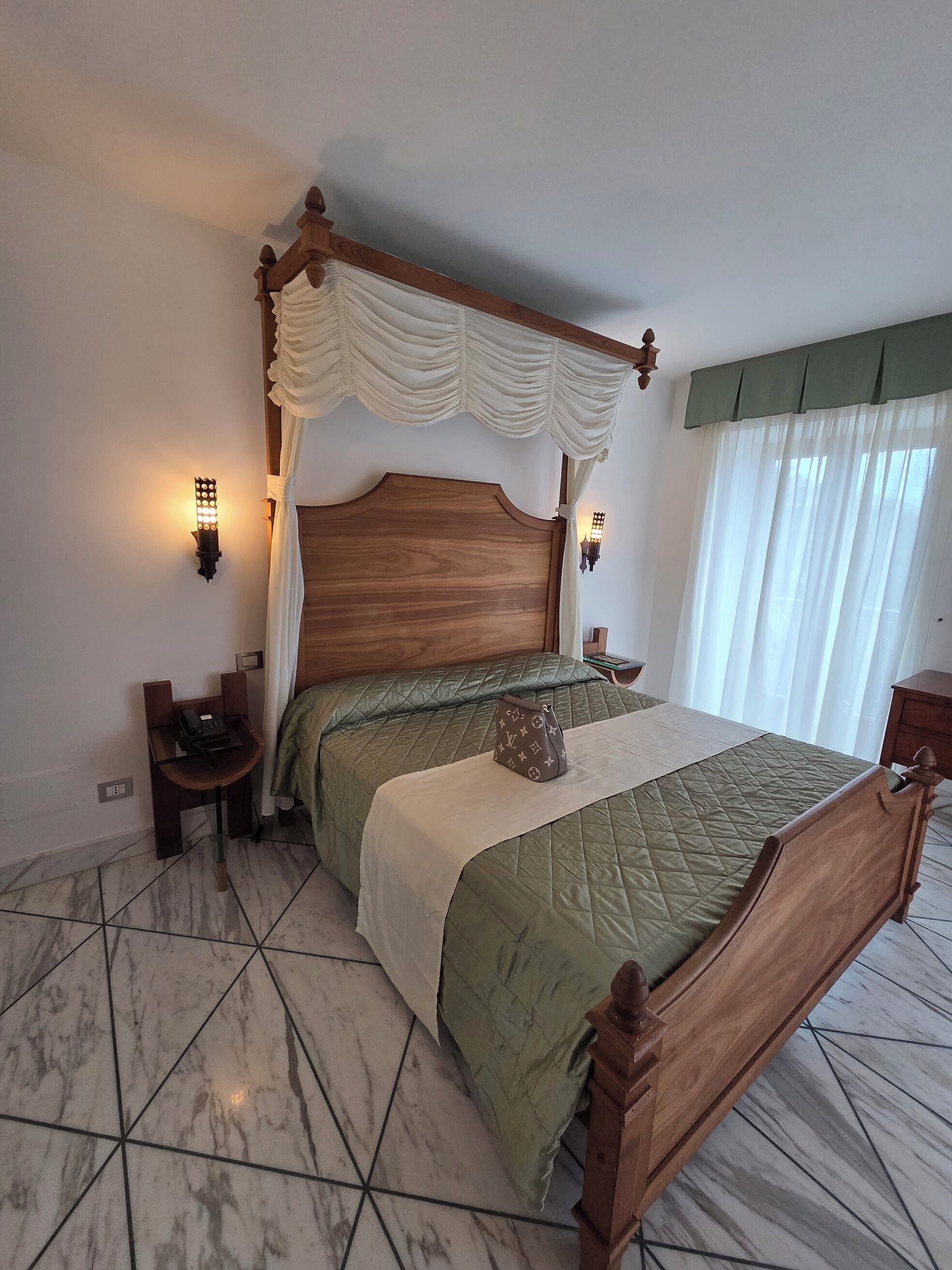 Een slaapkamer met een houten hemelbed, een groene sprei, een nachtkastje met een lamp en een bijzettafeltje met een telefoon.
