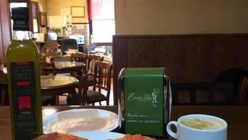 Una mesa de comedor con un plato de comida, una botella de aceite de oliva, una cafetera verde y una taza de café.