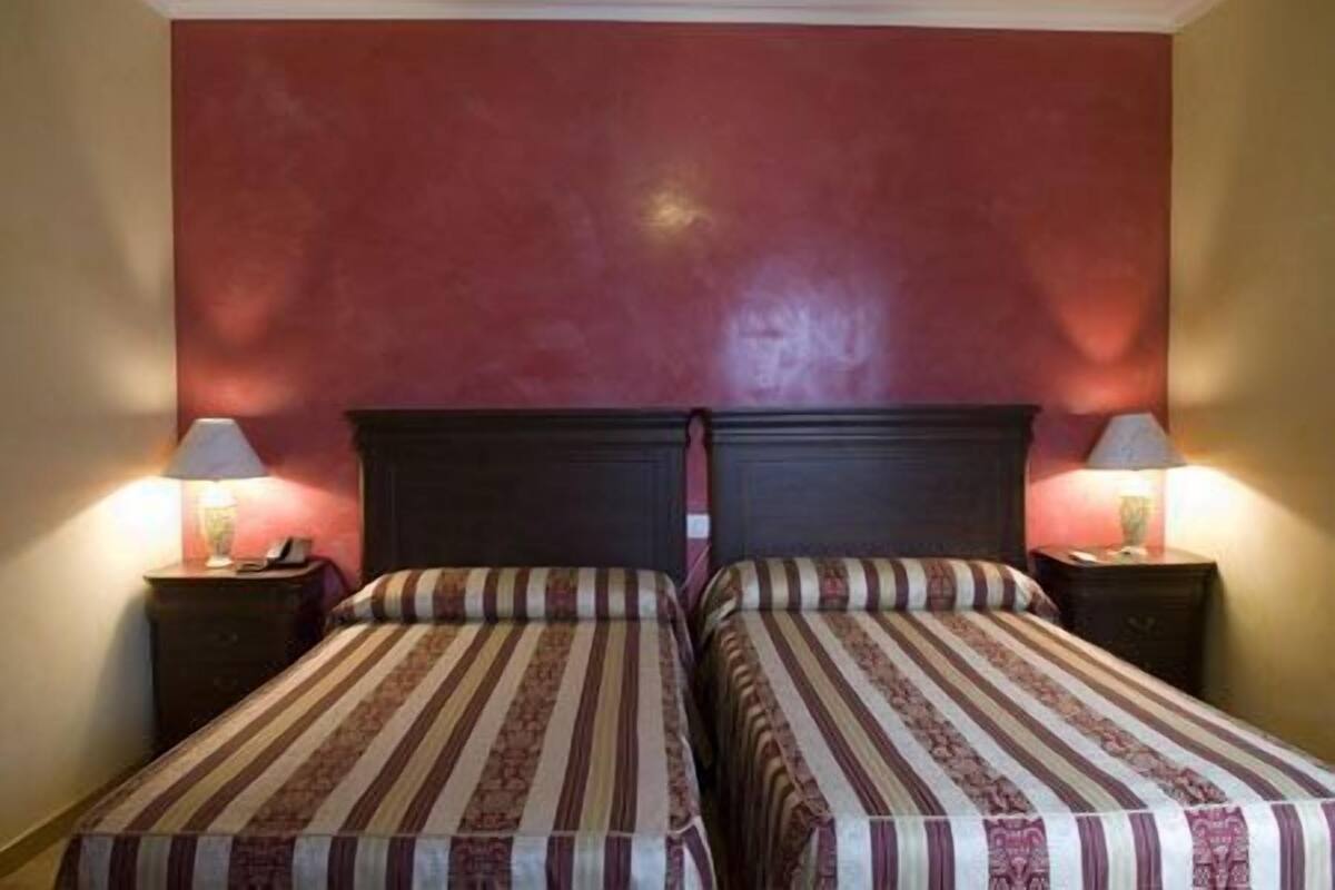 Ein Zimmer mit zwei Betten, jeweils mit gestreifter Bettdecke und dunklem Kopfteil. Auf jedem Nachttisch stehen eine Lampe und ein kleines dekoratives Objekt. Die Wände sind einfarbig gestrichen.