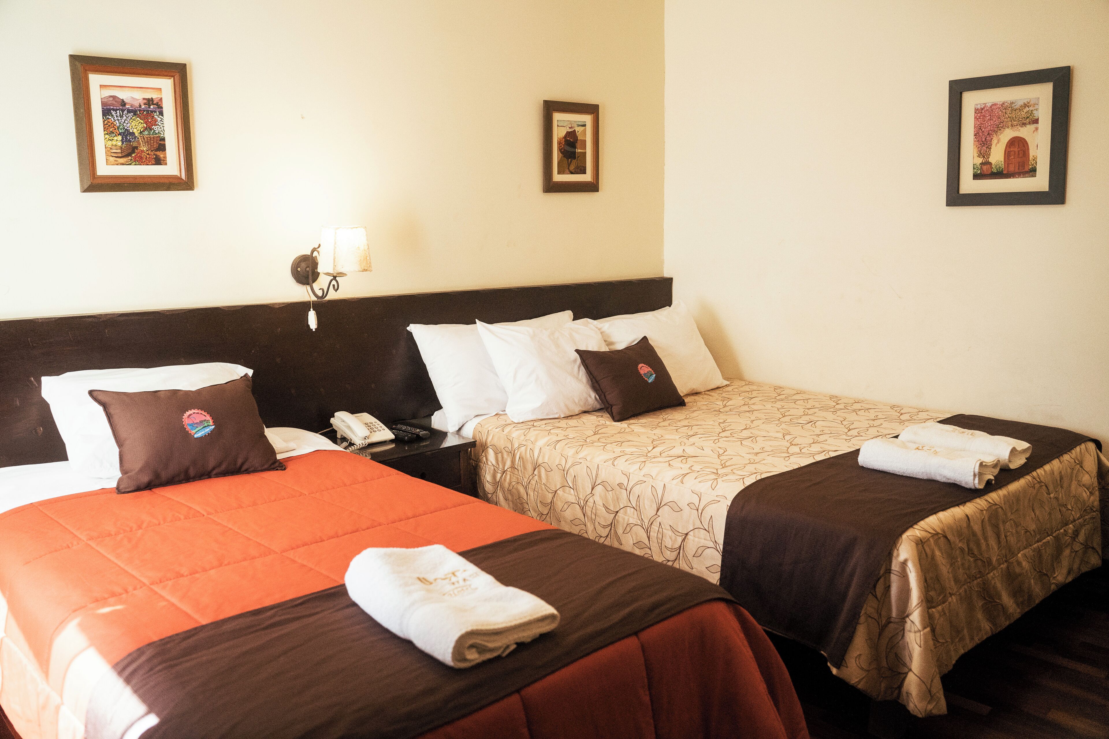 Habitación de hotel con dos camas, cada una con cubrecama marrón y almohadas blancas. En las paredes hay cuadros enmarcados, una mesita de noche con lámpara y un teléfono en la mesita.