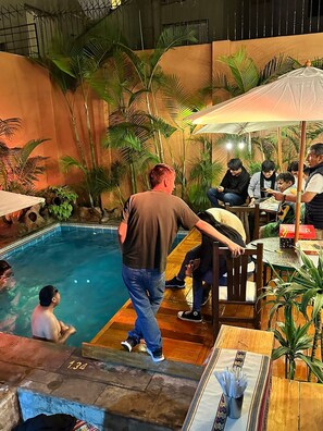 Zona de piscina con muelle de madera, rodeada de plantas tropicales y un área de estar cubierta con mesas y sombrillas.