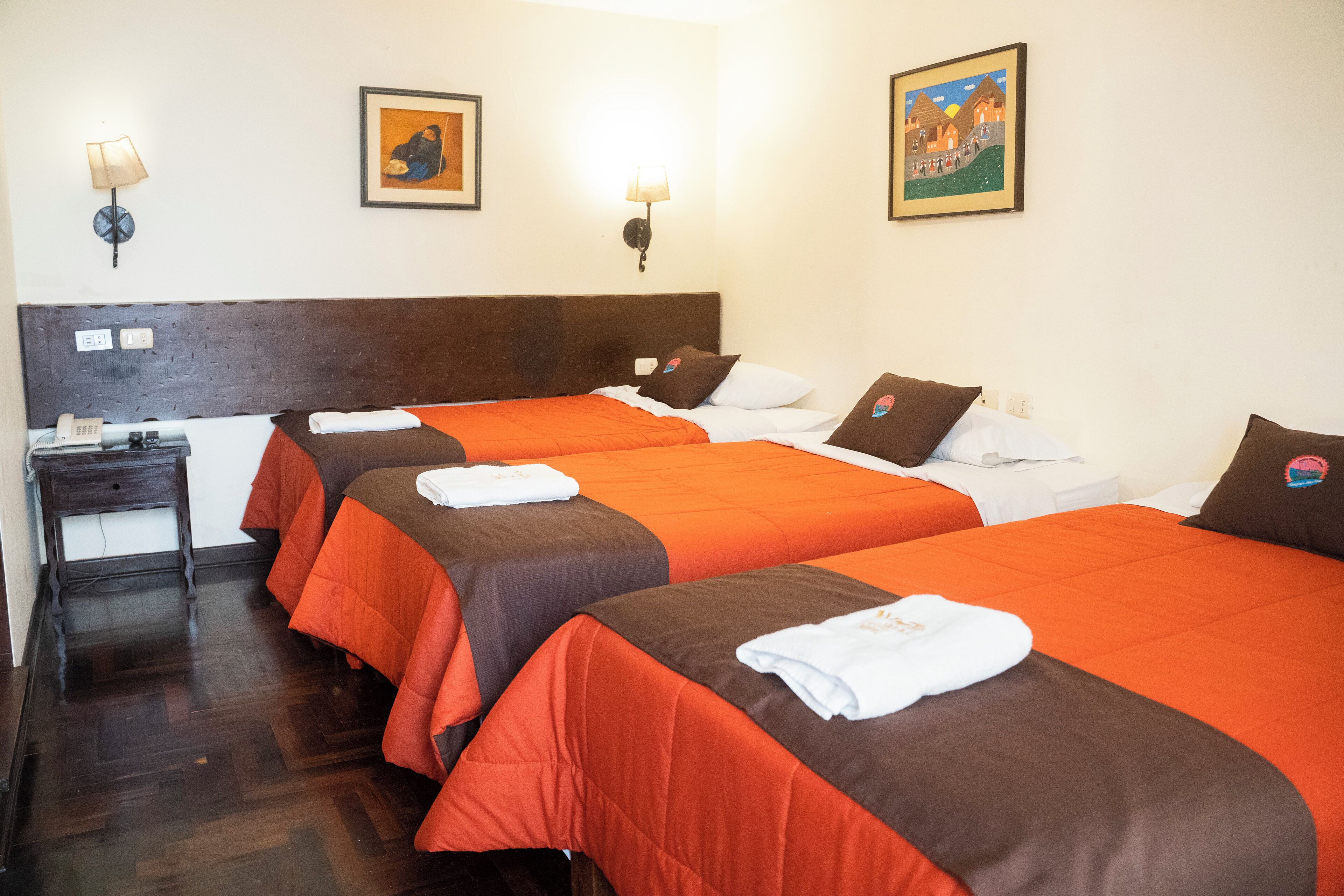 A hotel room with three beds, each with orange and brown bedding, a wooden headboard, a bedside table with a lamp, and two wall-mounted lamps.