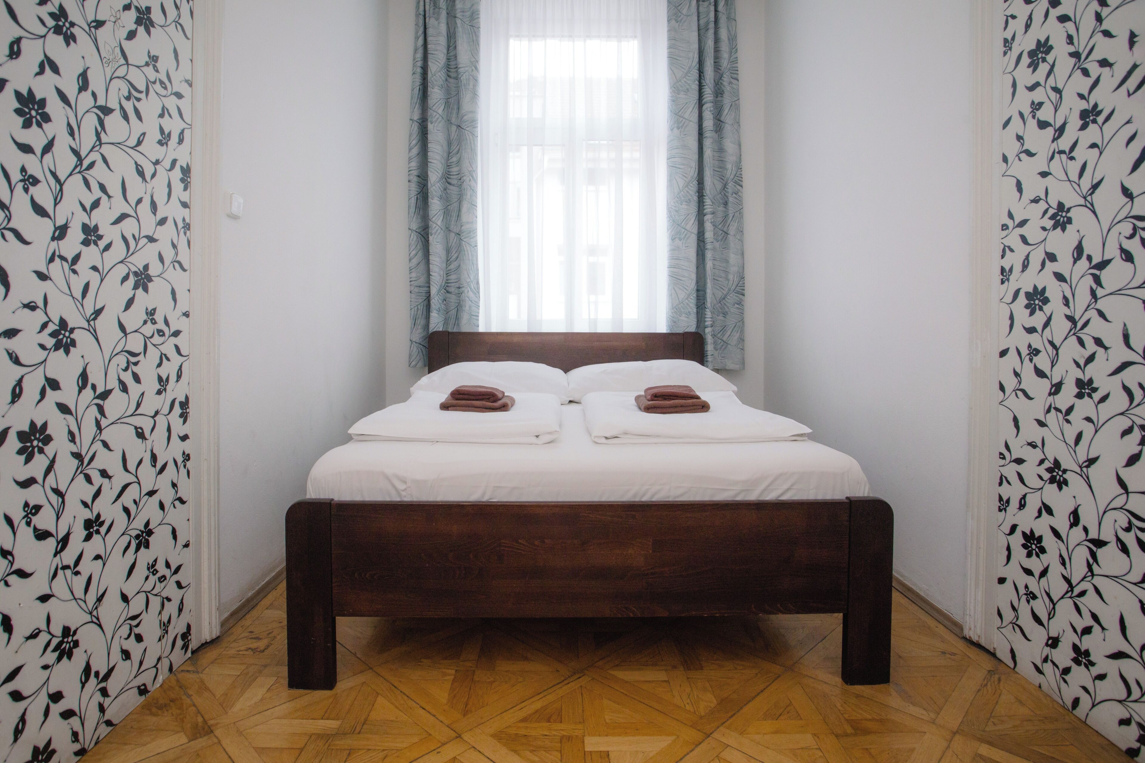 Uma cama de solteiro com roupa de cama branca e toalhas castanhas, uma cabeceira de madeira e uma janela com cortinas leves.