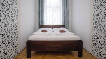 Uma cama de solteiro com roupa de cama branca e toalhas castanhas, uma cabeceira de madeira e uma janela com cortinas leves.