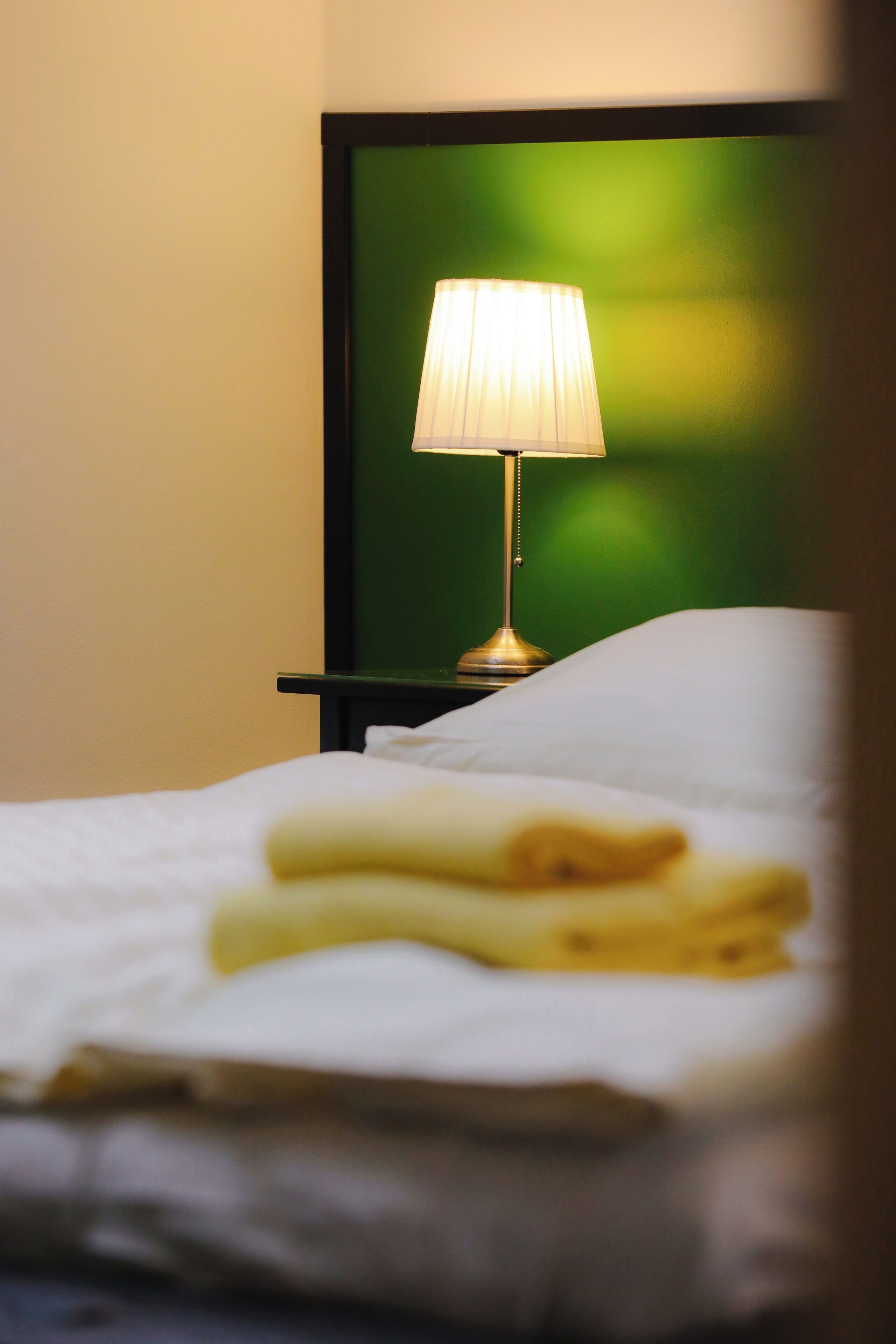 A neatly made bed with a folded white towel, a bedside table with a lamp, and a green wall in the background.