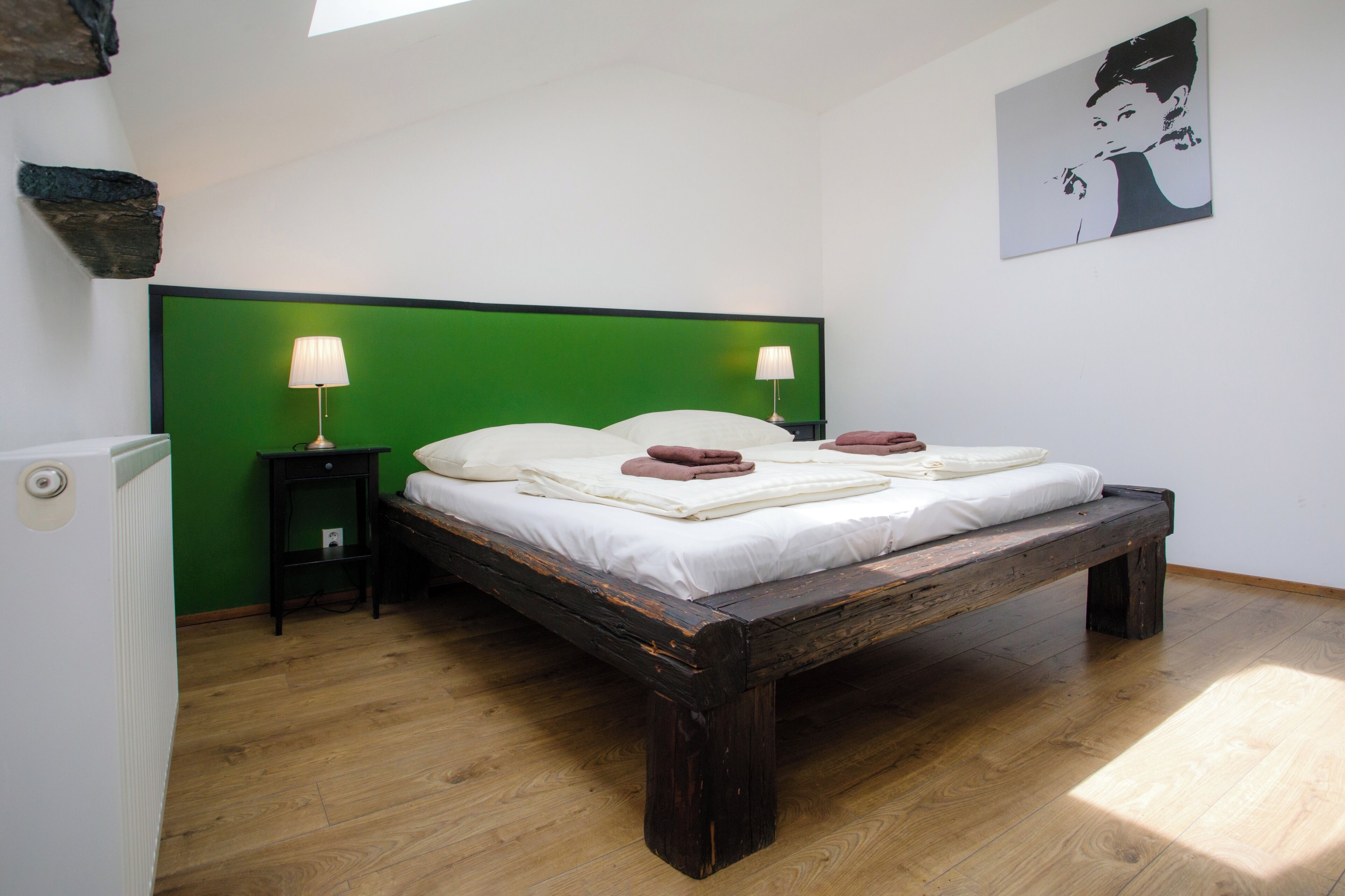 Un dormitorio con cabecera verde, estructura de cama de madera, dos mesitas de noche con lámparas y un cuadro colgado en la pared.