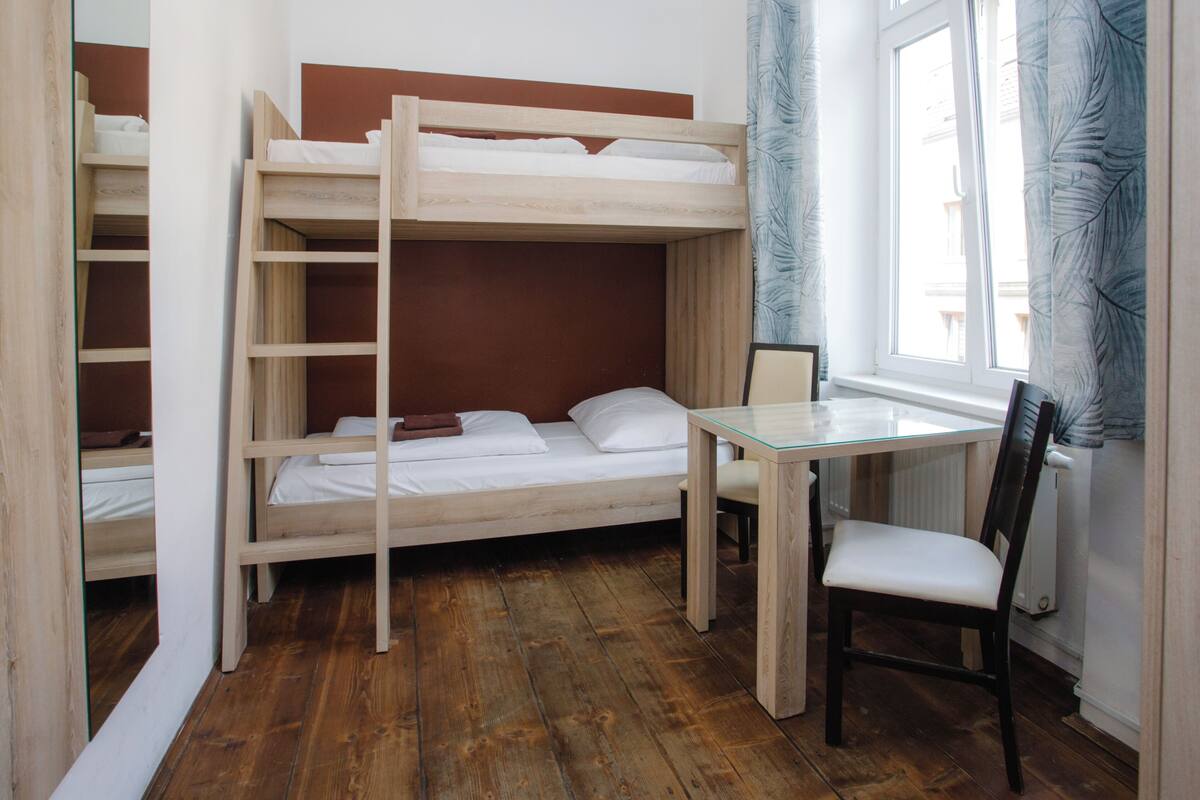 Ein Schlafraum mit Etagenbetten, einem Glastisch, einem Stuhl, Holzboden und einem Fenster mit Vorhängen.