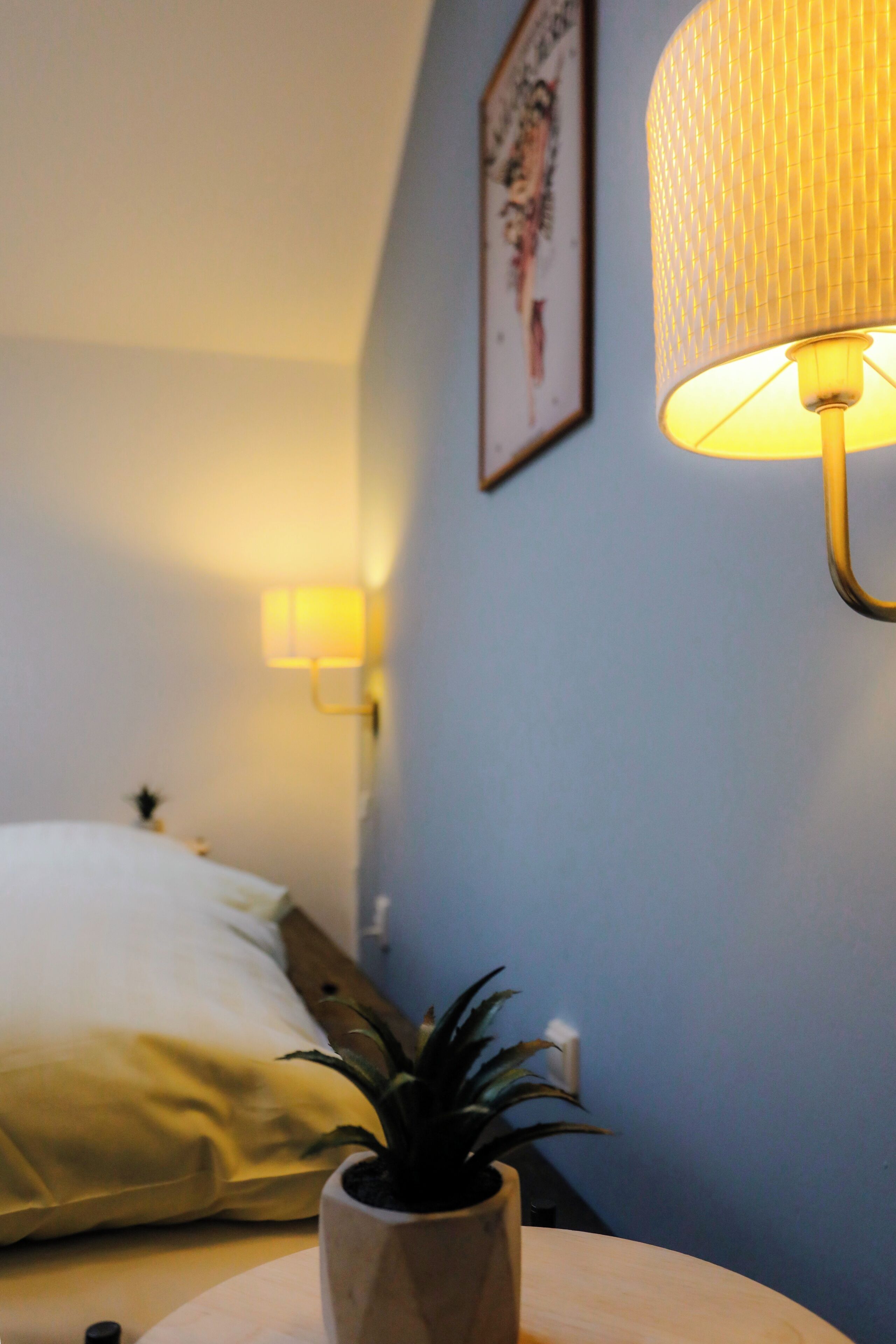 Una habitación con una cama, una lámpara de noche, una luz en la pared y una planta en maceta.