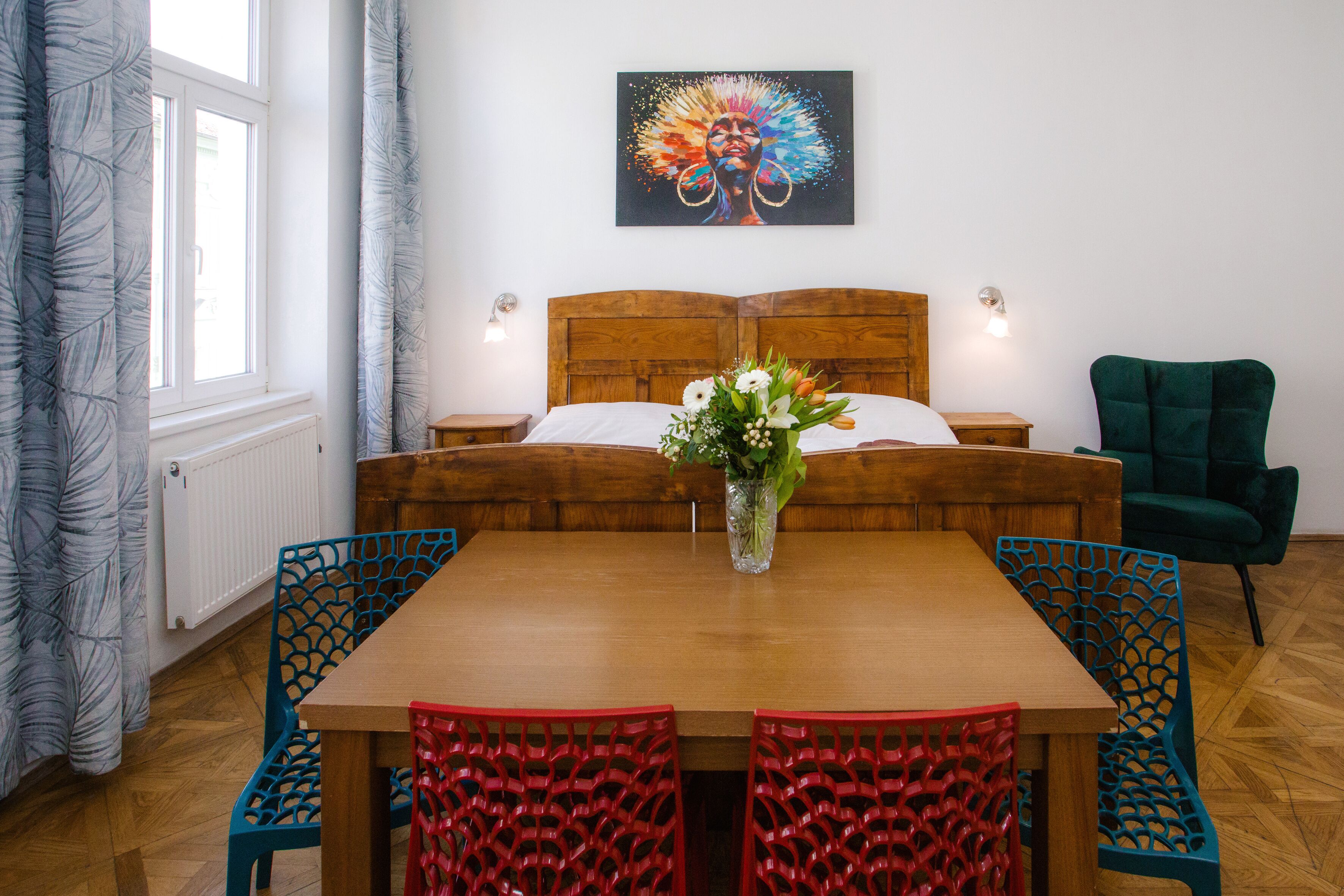 A hotel room with a wooden bed, a wooden table with chairs, a green armchair, and a painting on the wall.