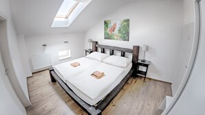Une chambre avec une lucarne, un lit recouvert de draps blancs, une tête de lit en bois, une table de chevet, une lampe et un tableau aux motifs tropicaux accroché au mur.