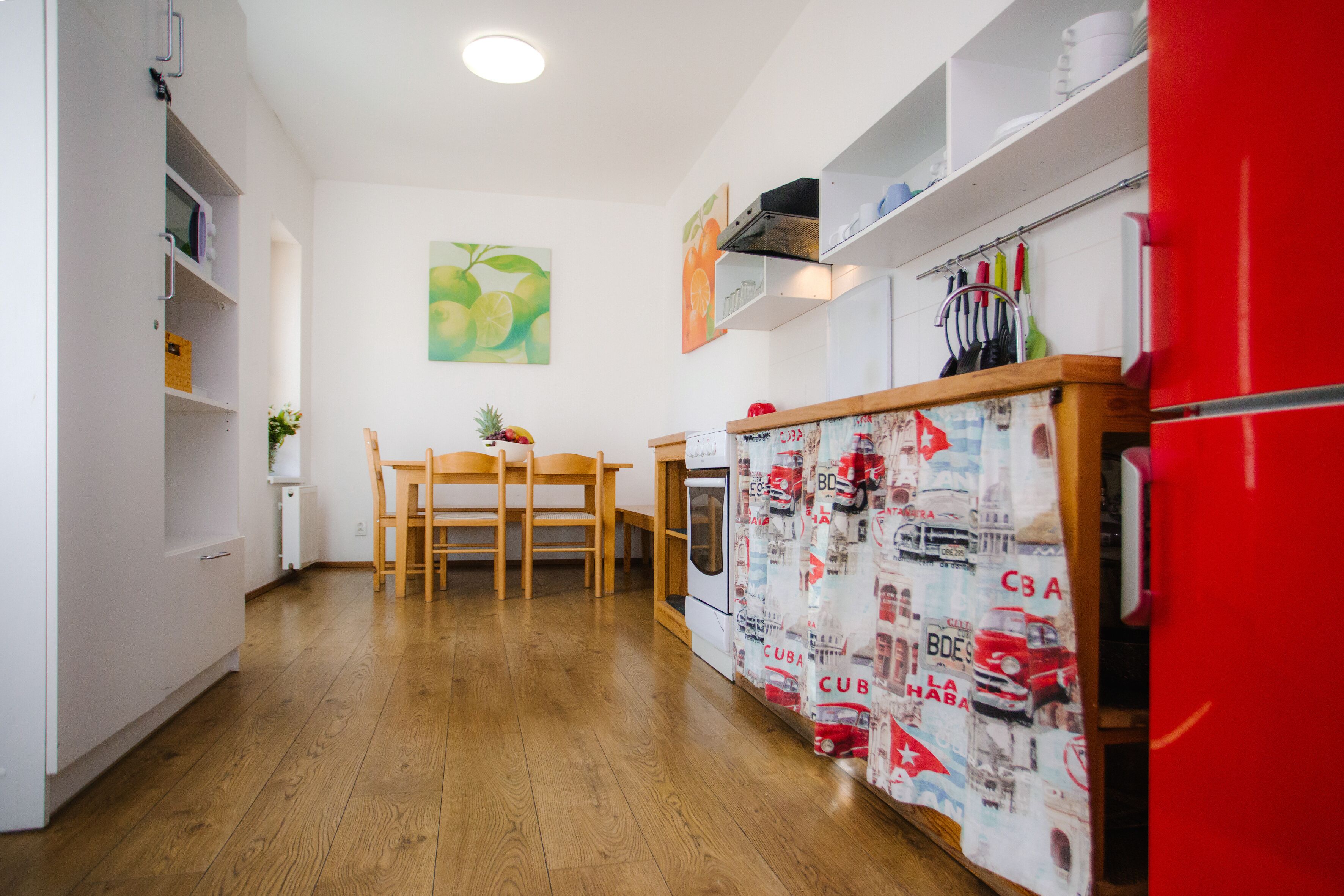 A kitchen with wooden flooring, a dining table with chairs, a refrigerator, and a wall with a fruit painting.