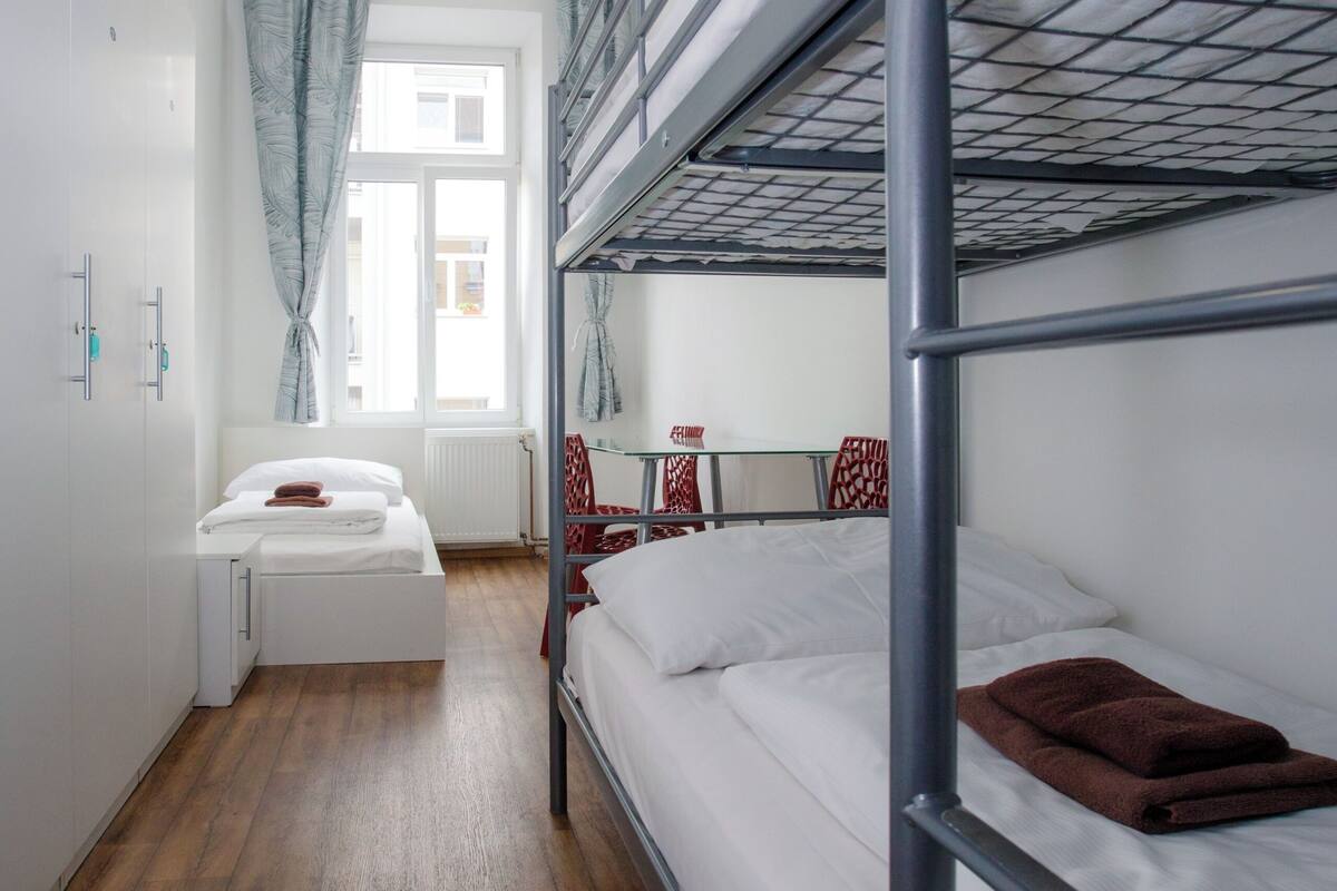 Ein Schlafsaalzimmer mit Etagenbetten, einem Fenster mit Vorhängen, einem Heizkörper und einem kleinen Tisch mit Stuhl.