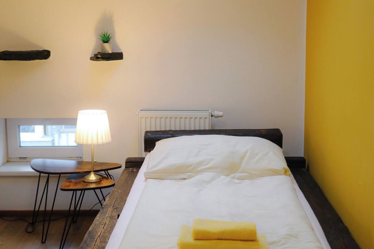 Ein Zimmer mit einem Bett, einem kleinen Tisch mit Lampe, einem Fenster und einer Heizung.