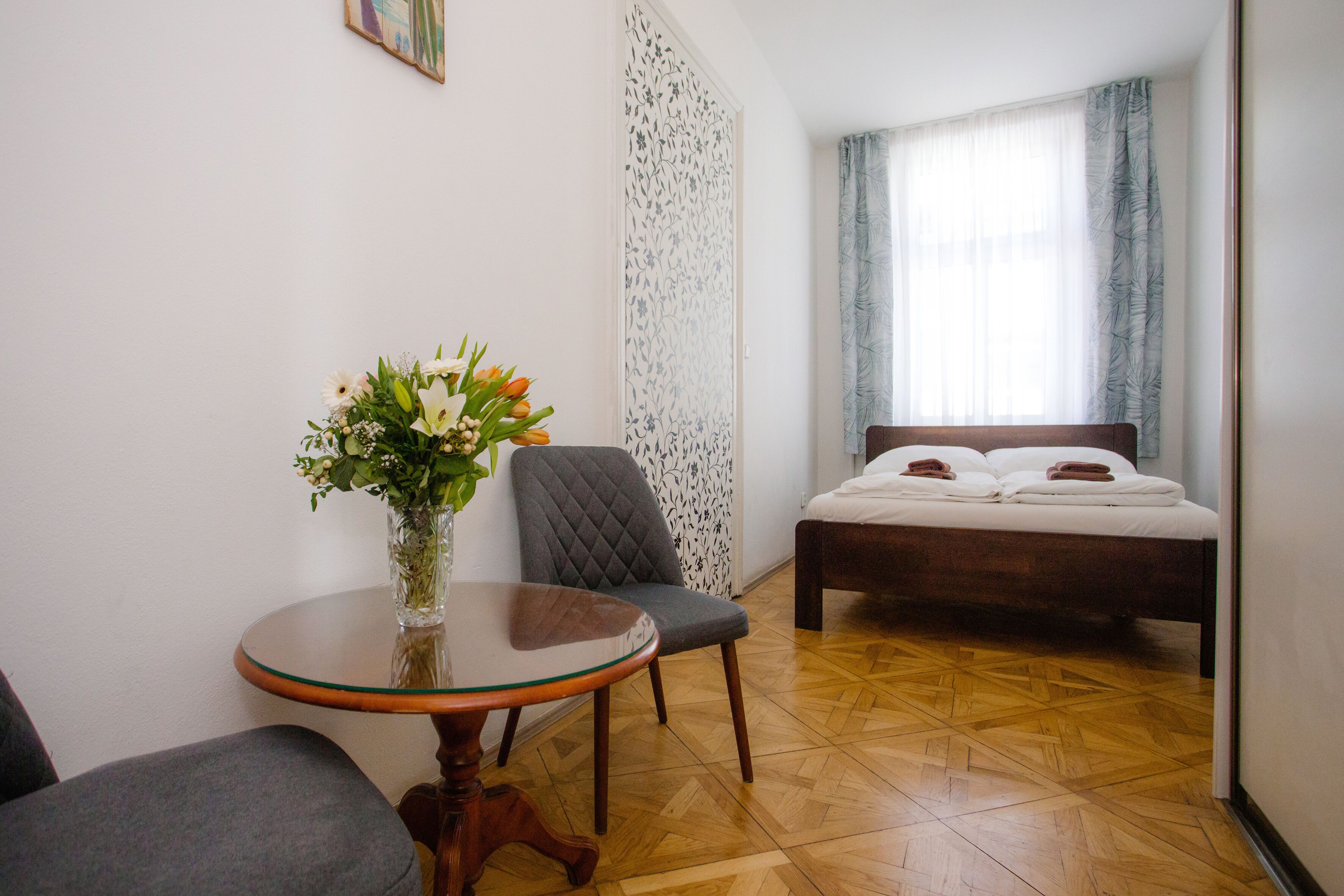 Ein Schlafzimmer mit Bett, Sessel, Tisch und einer Blumenvase, sowie einer Wand mit Muster.