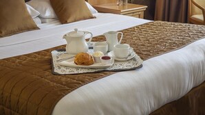 Una cama bien hecha con un edredón marrón, una bandeja con tetera y pasteles, y una mesita de noche de madera con una botella y una copa.